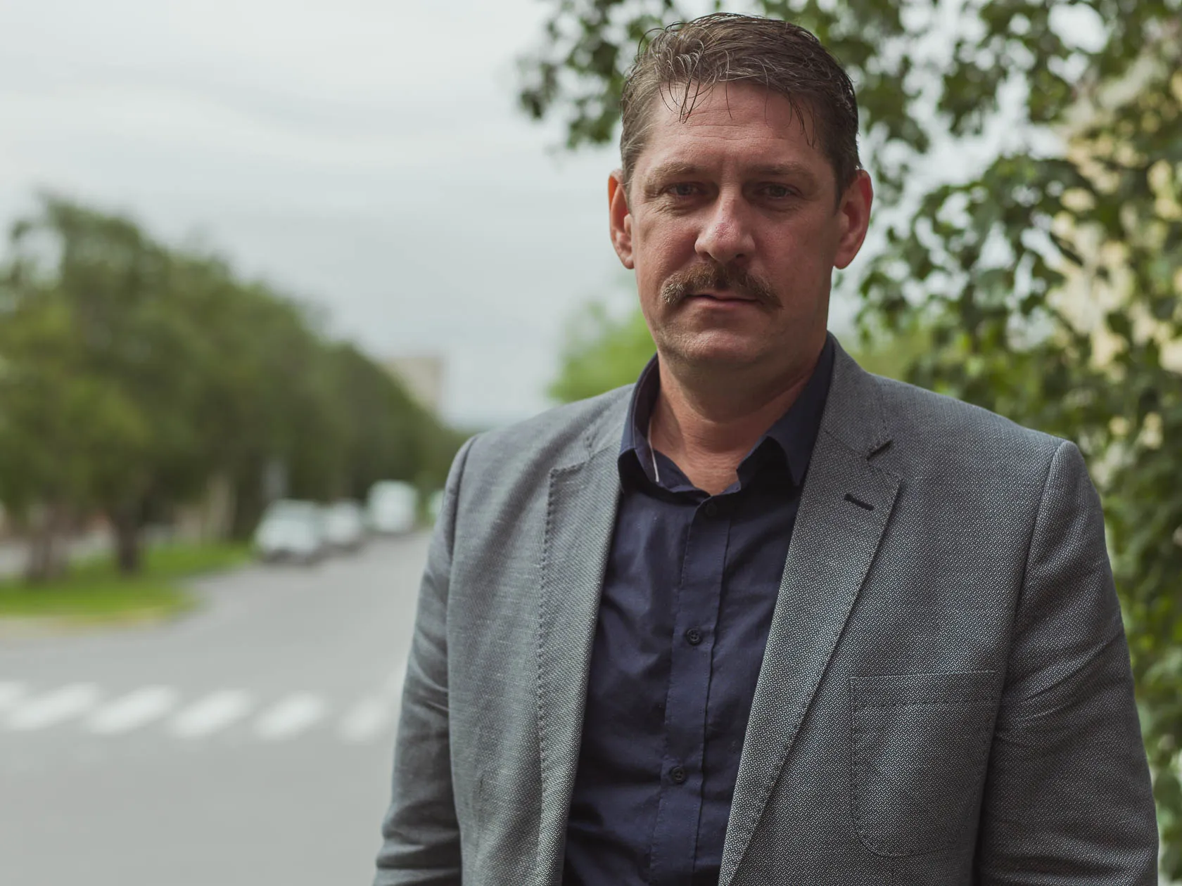 Мужчина с усами в сером пиджаке на фоне зелёной городской улицы