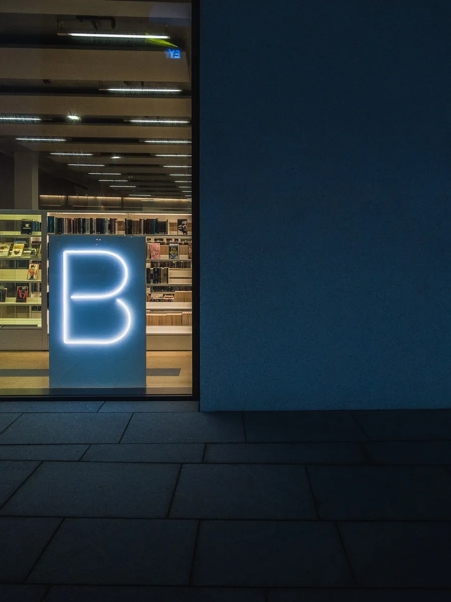 Светящаяся неоновая буква B у витрины библиотеки с книжными полками